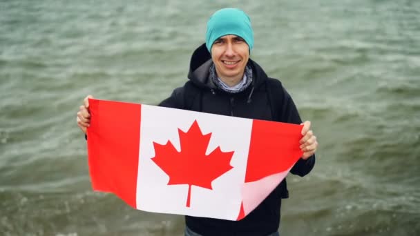 Slow motion portrait of joyful young man traveller holding Canadian flag in hands and looking at camera with glad smile. Sea waves are visible in background. — Stock Video