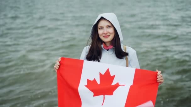 Retrato en cámara lenta de la alegre joven ciudadana orgullosa sosteniendo la bandera canadiense ondeando y sonriendo mirando a la cámara de pie cerca del océano . — Vídeos de Stock