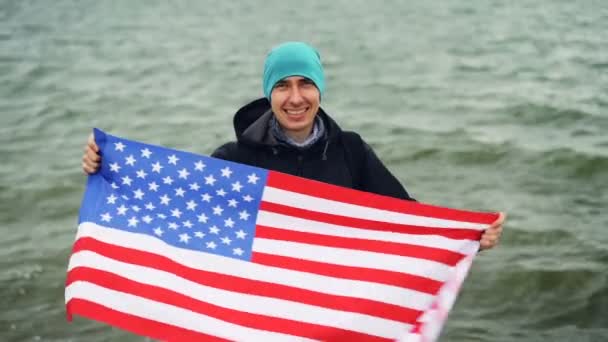 Zeitlupenporträt eines fröhlichen Reisenden, der die amerikanische Nationalflagge in Händen hält und sie bewegt, um die Freiheit zu feiern. Im Hintergrund sind schöne Meereswellen zu sehen. — Stockvideo