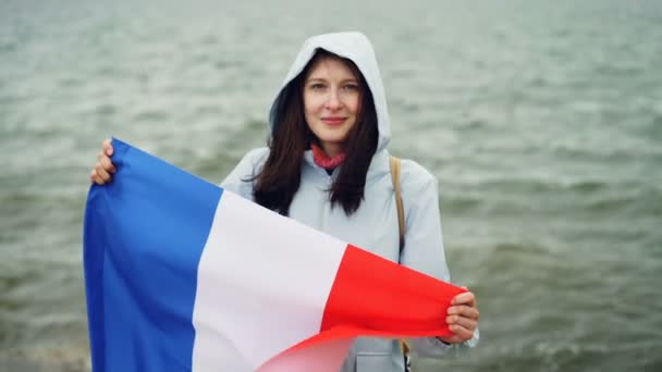 Retrato en cámara lenta del alegre patriota francés sosteniendo la bandera oficial de Francia con una linda sonrisa de pie cerca del agua y sonriendo. Concepto de nacionalismo y juventud . — Vídeos de Stock