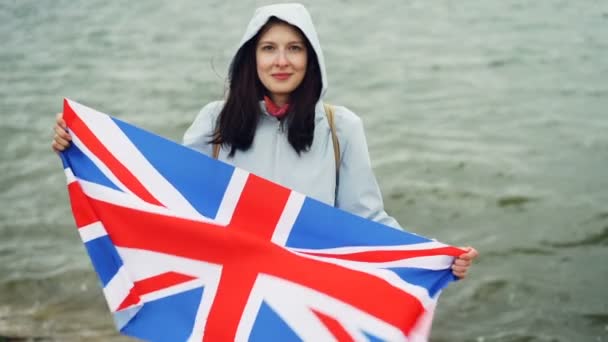 Slow motion portrait of glad tourist visiting Great Britain holding British national flag and smiling standing on sea shore with beautiful nature visible. — Stock Video