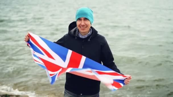 Slow motion portrait of patriotic Englishman holding flag of Great Britain standing on sea coast and smiling happily. Proud citizens, foreign countries and people concept. — Stock Video