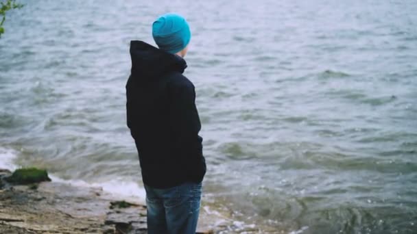 Slow motion portrait of thoughtful guy in hat, jacket and jeans standing on the sea coast and watching water waves enjoying nature. Healthy lifestye and tourism concept. — Stock Video
