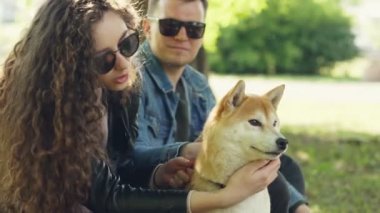Mutlu çift kız ve erkek arkadaşı parkta şirin köpek yavrusu okşamaya ve konuşuyor. Kadın ve adam rahat kıyafetler ve güneş gözlüğü takıyorsun, güzel park arka planda.