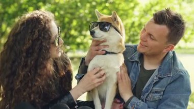 Gülümseyen köpek sahipleri gözleri onun üzerinde güneş gözlüğü ile köpek yavrusu okşamaya ve gülümseyen güneşli yaz günü parkta dinlenirken. Eğlenceli, Evcil hayvanlar ve insanlar kavramı.