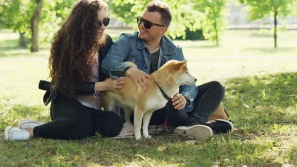 Ευτυχείς φίλοι κορίτσι και ο τύπος χαϊδεύει αξιολάτρευτο σκυλί, μιλώντας και χαμογελώντας, ενώ κάθονται στο πάρκο μαζί φορώντας γυαλιά ηλίου. Φύση, άνθρωποι και ζώα έννοια. — Αρχείο Βίντεο