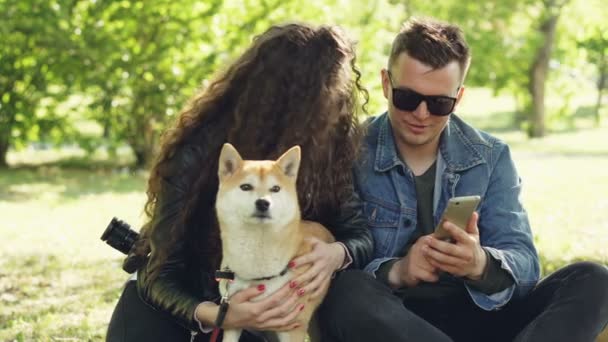 Ung kvinna är petting sin hund som sitter på gräset medan hennes pojkvän visar hennes smartphone skärmen, människor pratar och skrattar. Helgen i begreppet park. — Stockvideo