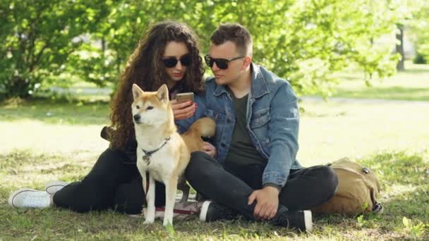 Mari et femme utilisent smartphone reposant sur la pelouse dans le parc, regarder l'écran et parler et chien agité. Technologie moderne, animaux domestiques et concept de ville . — Video