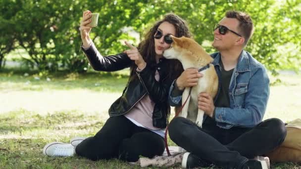 Ευτυχισμένο ζευγάρι είναι η λήψη selfie με το σκυλί τους, χρησιμοποιώντας το smartphone κάθεται στο γρασίδι στο πάρκο, άτομα που παρουσιάζουν, μιλάμε και χάιδεμα shiba inu κουτάβι. — Αρχείο Βίντεο