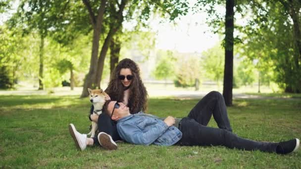 Bello giovane uomo è sdraiato sull'erba nel parco, mentre la sua ragazza sta parlando con lui e accarezzare cane carino, coppia sta ridendo e godendo fine settimana . — Video Stock