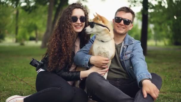 Πορτρέτο του χαριτωμένο ζευγάρι και shiba inu σκυλί όλα φορώντας γυαλιά ηλίου βλέπουν φωτογραφική μηχανή και χαμογελαστός κάθεται στο γρασίδι στο πάρκο. Σύγχρονος τρόπος ζωής, χαριτωμένα ζώα και φύση της έννοιας. — Αρχείο Βίντεο