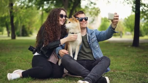 Handsome young man is taking selfie with his pretty wife and cute dog, all wearing sunglasses. Guy is holding smartphone taking pictures and posing. — Stock Video