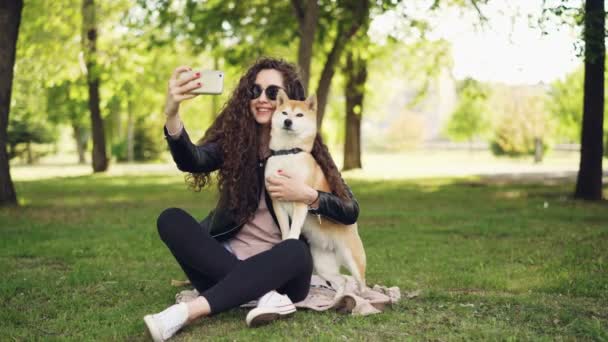 陽気なブロガーのペット所有者を取っている selfie のスマート フォンを使用して彼女の犬と人間と動物は公園の芝生の上に座っているとポーズ、女性は caressing し犬のキス. — ストック動画