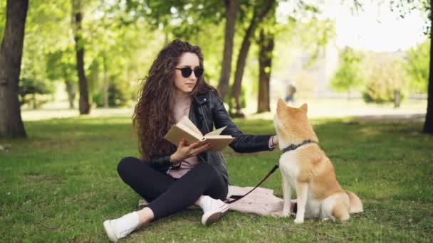 Glada kvinnlig student är läsebok i parken sitter på gräsmattan och smekande vacker hund, sällskapsdjur gäspningar och slicka sin munkorg, tjej skrattar. — Stockvideo