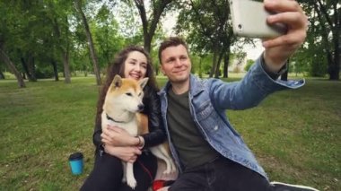 Mutlu çift park öpüşme içinde selfie alarak ve genç bir kadın tutarak ve şirin köpek yavrusu shiba Inu doğurmak okşama süre smartphone vasıl arıyorsunuz.
