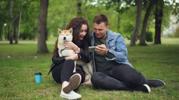 Loving boyfriend is showing smartphone screen to pretty young lady who is holding and patting her well-bred dog while sitting on lawn in city park. People and technology concept. — Stock Video