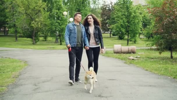 Веселый парень в джинсовой одежде выгуливает собаку, держа жену за руку, летом люди и животные идут по дорожке в парке. Концепция активного образа жизни . — стоковое видео