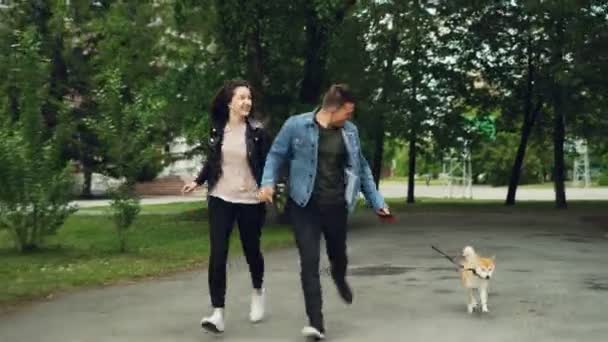 Улыбающиеся молодые люди муж и жена бегают по парку с милой собакой, наслаждаясь активностью и держась за руки. Романтические отношения, любящие животные и концепция физических упражнений . — стоковое видео