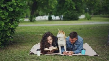 Genç Aile hafta sonu harcama park, kadın okuma kitabı, smartphone, sevimli köpek aralarında dinlenme kullanan adam. Adam karısına telefon ekranını göstermeyi.