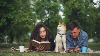 Mutlu çiftimiz parkta dinleniyor, kadın kitap okurken erkek smartphone kullanarak, onların köpek battaniye aralarında oturuyor. İnsanlar konuşmak ve gülümseyen.