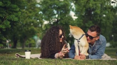 Genç kadın komik resimler Smartphone'da sevgilisi ile evde beslenen hayvan köpek parkta dinlenirken gösteriyor, insanlar ekose çim ve izlerken ekran üzerinde yalan.