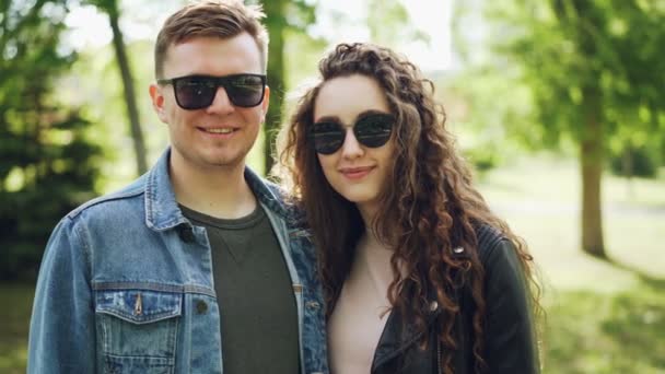 Portrét šťastnému páru muž a žena nosí sluneční brýle a módní oblečení, při pohledu na fotoaparát a usmívá se s krásným zeleným parkem v pozadí. — Stock video