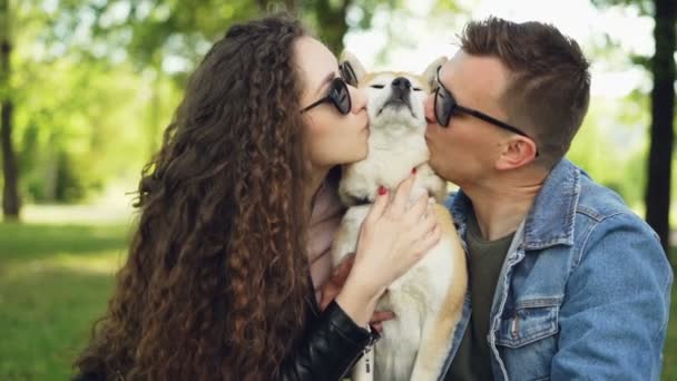 Zwolnionym tempie szczęśliwa para Całowanie piękny pies, śmiejąc się i rozmawiając, siedząc na trawie w parku, ciesząc się latem. Kochający zwierzęta, relacji i koncepcja wypoczynek. — Wideo stockowe