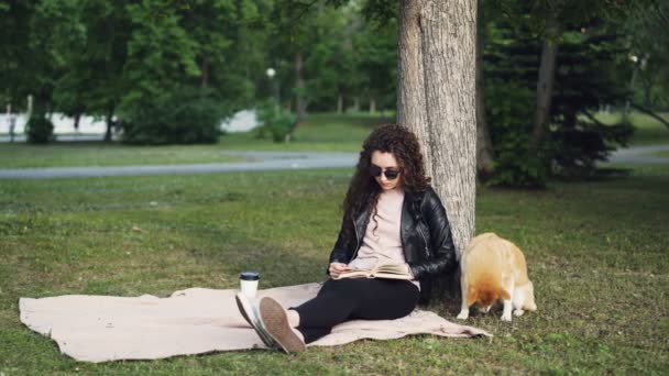 放松的女孩是看书和喝外卖咖啡坐在公园的草坪上, 而她可爱的狗是闻地面和空气坐在树下。休闲和宠物概念. — 图库视频影像