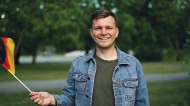 Gülümseyen ve kameraya bakarak Almanya, resmi bayrağı sallayarak neşeli Alman turist ağır çekim portre. Güzel park arka plan, sıcak yaz günü olduğunu..