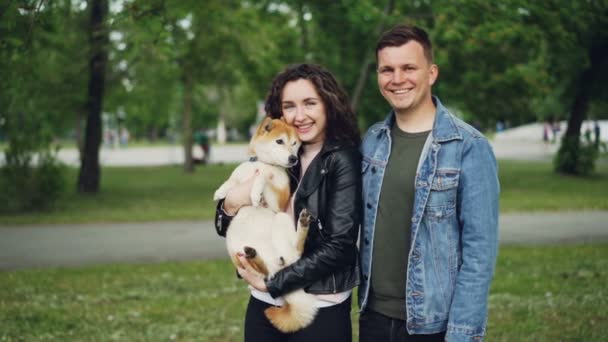 Slow motion portrait of attractive young woman brunette holding her dog standing in the park with boyfriend. Happy people are looking at camera and smiling. — Stock Video