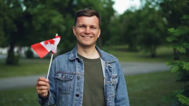 Slow motion portrait of handsome guy in casual clothes waving Canadian flag, smiling and looking at camera standing in beautiful park. People and patriotism concept. — Stock Video