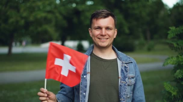 Retrato en cámara lenta de los fanáticos de los deportes suizos sonriendo, ondeando la bandera de Suiza y mirando a la cámara con árboles verdes y césped en el fondo. Concepto juventud y nacionalismo . — Vídeos de Stock