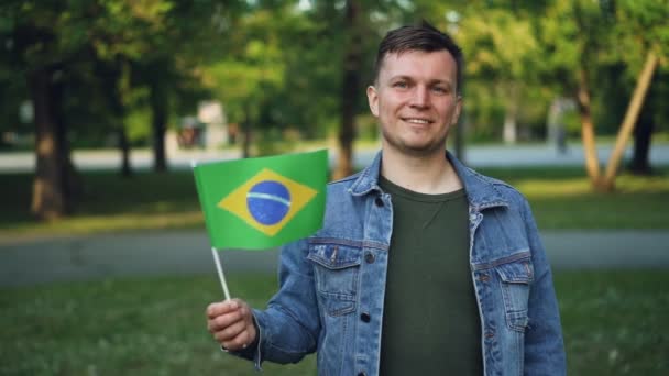Медленный кинопортрет счастливого путешественника эмоционального молодого человека, размахивающего бразильским флагом после посещения латиноамериканской страны. Человек и туризм . — стоковое видео