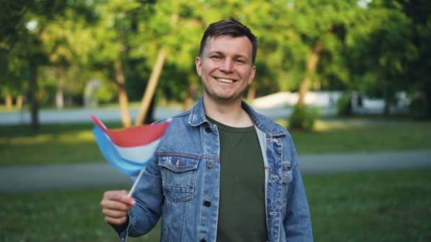 Retrato en cámara lenta del alegre tipo feliz turista ondeando la bandera de los Países Bajos, sonriendo y mirando a la cámara. Orgullo nacional, visita a países y concepto de personas . — Vídeos de Stock