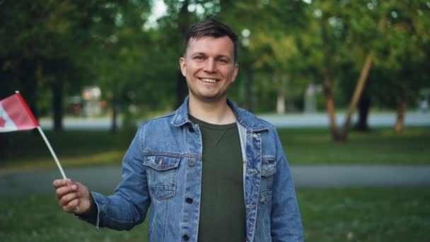 Retrato em câmera lenta de um jovem atraente fã canadense de esportes acenando com a bandeira do Canadá apoiando a equipe esportiva nacional, sorrindo e olhando para a câmera . — Vídeo de Stock