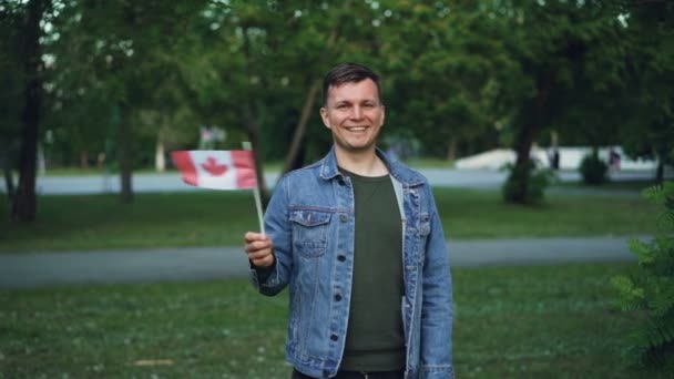 Slow motion portret van knappe mannelijke reiziger vrolijke kerel Canadese vlag zwaaien, camera kijken en glimlachend staande in het prachtige stadspark met bomen en gazons. — Stockvideo