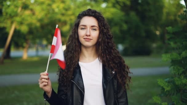 Retrato en cámara lenta de la alegre joven austríaca ondeando la bandera de Austria, mirando a la cámara y sonriendo de pie en el parque con grees en el fondo . — Vídeos de Stock