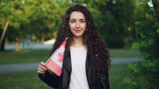 Retrato en cámara lenta de un feliz fanático de los deportes canadienses ondeando la bandera nacional de Canadá mirando a la cámara con una sonrisa alegre mientras está parado afuera en el parque en el día de verano . — Vídeos de Stock