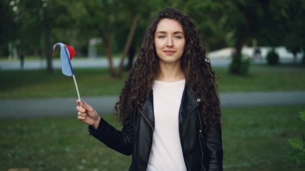 Slow motion portrait of French woman patriot waving flag of France with glad smile and looking at camera. Beautiful green park with trees and lawns is in background. — Stock Video