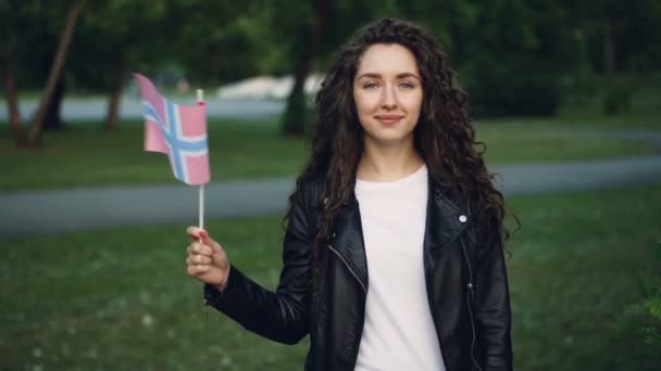 Slow motion portrait of world traveller happy girl waving Norwegian flag, looking at camera and smiling. Beautiful park with green trees and nice lawns is visible. — Stock Video