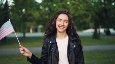 Amerika Birleşik Devletleri bayrağı sallayarak ve gülümseyerek kameraya bakarak mutlu Amerikalı turist ağır çekim portre. Seyahat, yabancı ülkeler ve insanlar kavramı ziyaret dünya.