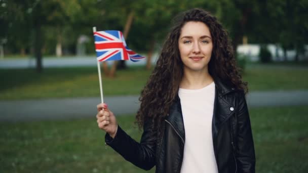 Медленный портрет счастливого британского студента, размахивающего флагом Великобритании, смотрящего в камеру и улыбающегося, стоящего на улице в парке. Концепция людей и стран . — стоковое видео