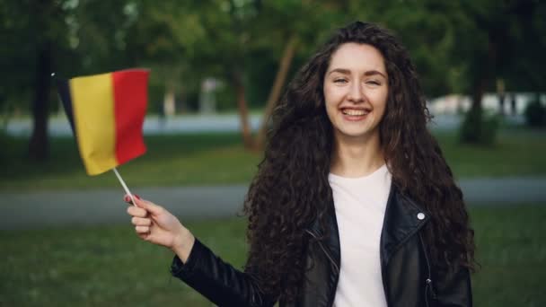 Медленный портрет привлекательной девушки-путешественницы, размахивающей бельгийским флагом, смотрящей в камеру и улыбающейся, стоящей летом в зеленом парке. Человек и туризм . — стоковое видео