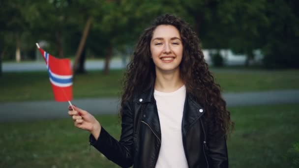 Retrato en cámara lenta de una dama alegre con ropa casual ondeando bandera noruega, mirando a la cámara y sonriendo. Gente feliz, cuntries europeos y concepto de viaje . — Vídeo de stock