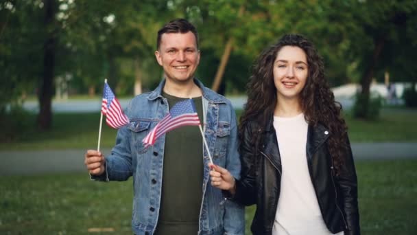 Retrato en cámara lenta de una feliz pareja estadounidense ondeando banderas estadounidenses paradas en el parque juntas, mirando a la cámara y sonriendo. Concepto de relación, personas y países . — Vídeo de stock