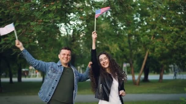 Retrato em câmera lenta de fãs de esportes americanos homem e mulher acenando bandeiras dos EUA enquanto pulava e ria no parque da cidade olhando para a câmera. Felicidade e conceito de pessoas . — Vídeo de Stock