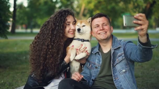 Câmera lenta de menina par feliz e cara tomando selfie com cão bonito posando e beijando animal segurando smartphone. Tecnologia moderna, parques e conceito de lazer . — Vídeo de Stock