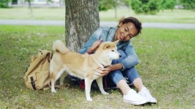 Oldukça karışık ırk kadın parkta çim üzerinde sıcak yaz günü dinlenme onu evde beslenen hayvan köpek mızmızlanıyor. Güzel yeşil çim, yaşlı ağaçlar ve sırt çantası görülebilir.