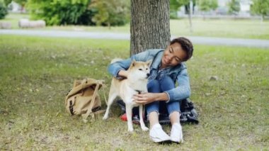 Genç Afrikalı-Amerikalı kadın sevimli köpek yavrusu parkta yeşil çimenlerin üzerinde dinlenme ve evde beslenen hayvan şefkat ve hayranlığı ile konuşmaya okşama. İnsanlar ve hayvanlar kavramı.