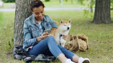 Mutlu Afrikalı-Amerikalı kadın smartphone kullanarak ve onun sevimli hayvan köpek şehir parkında rüzgarlı yaz günü dinlenme okşama. Doğa, hayvanlar ve insanlar kavramı.